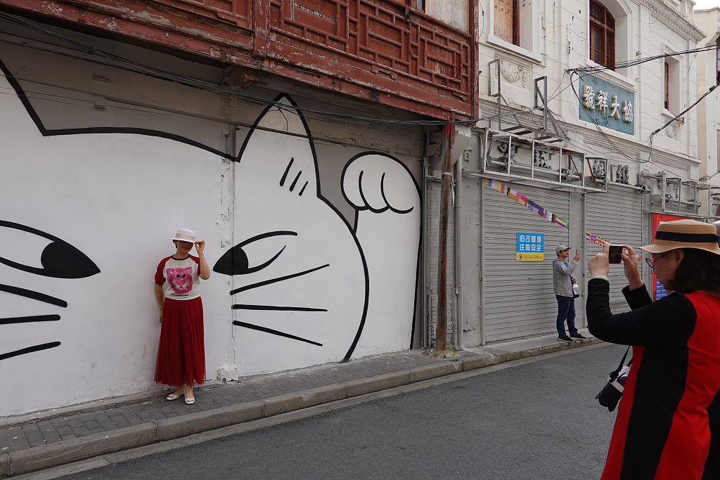 A woman poses for a photo with a cartoon cat image on a wall at the Fangbangzhonglu Road in Shanghai, May 25, 2023. /CFP