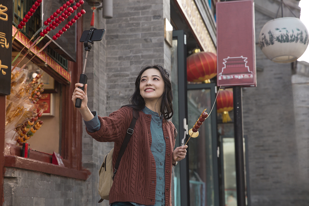 This file photo shows a young woman taking a selfie in Beijing's Qianmen district with a local tanghulu snack in hand.  Made with sweet red hawthorn berries, the stick candy is an iconic Beijing street snack.  /CFP