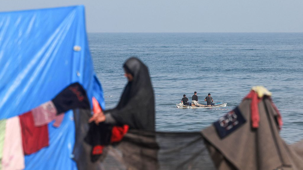 A displaced Palestinian woman hangs clothes to dry near her tent as fishermen sail in a boat in Rafah, southern Gaza, March 30, 2024. /CFP