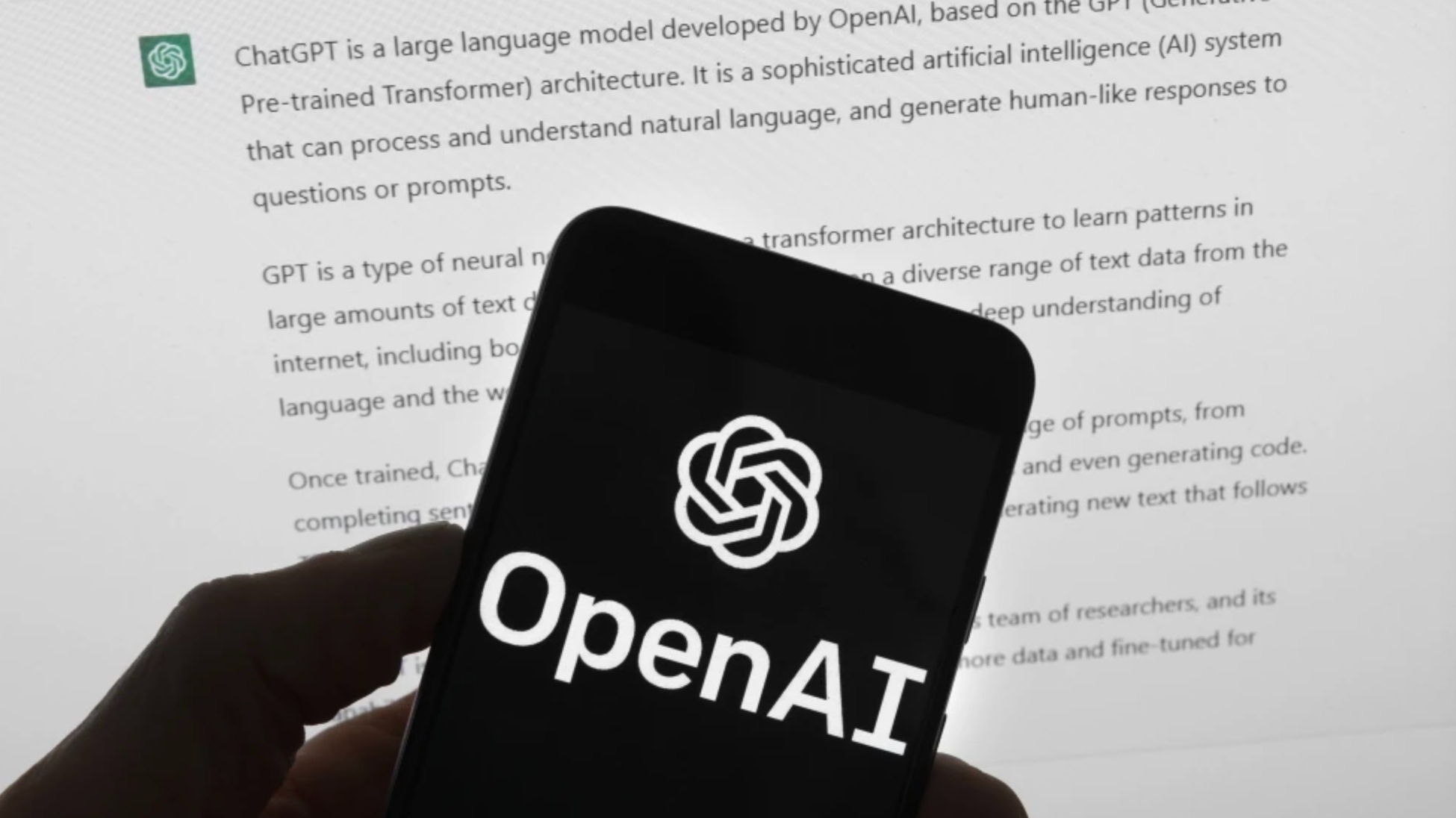 The OpenAI logo is seen on a mobile phone in front of a computer screen displaying output from ChatGPT. /AP