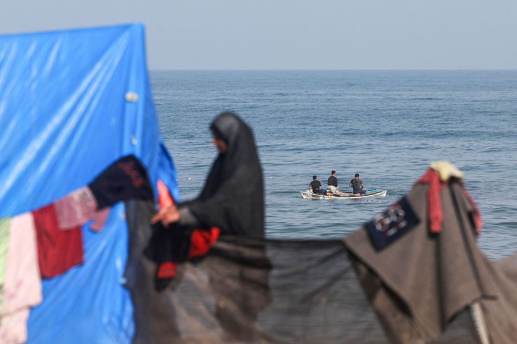 A displaced Palestinian woman hangs clothes to dry near her tent as fishermen sail in a boat in Rafah in the southern Gaza Strip, March 30, 2024. /CFP