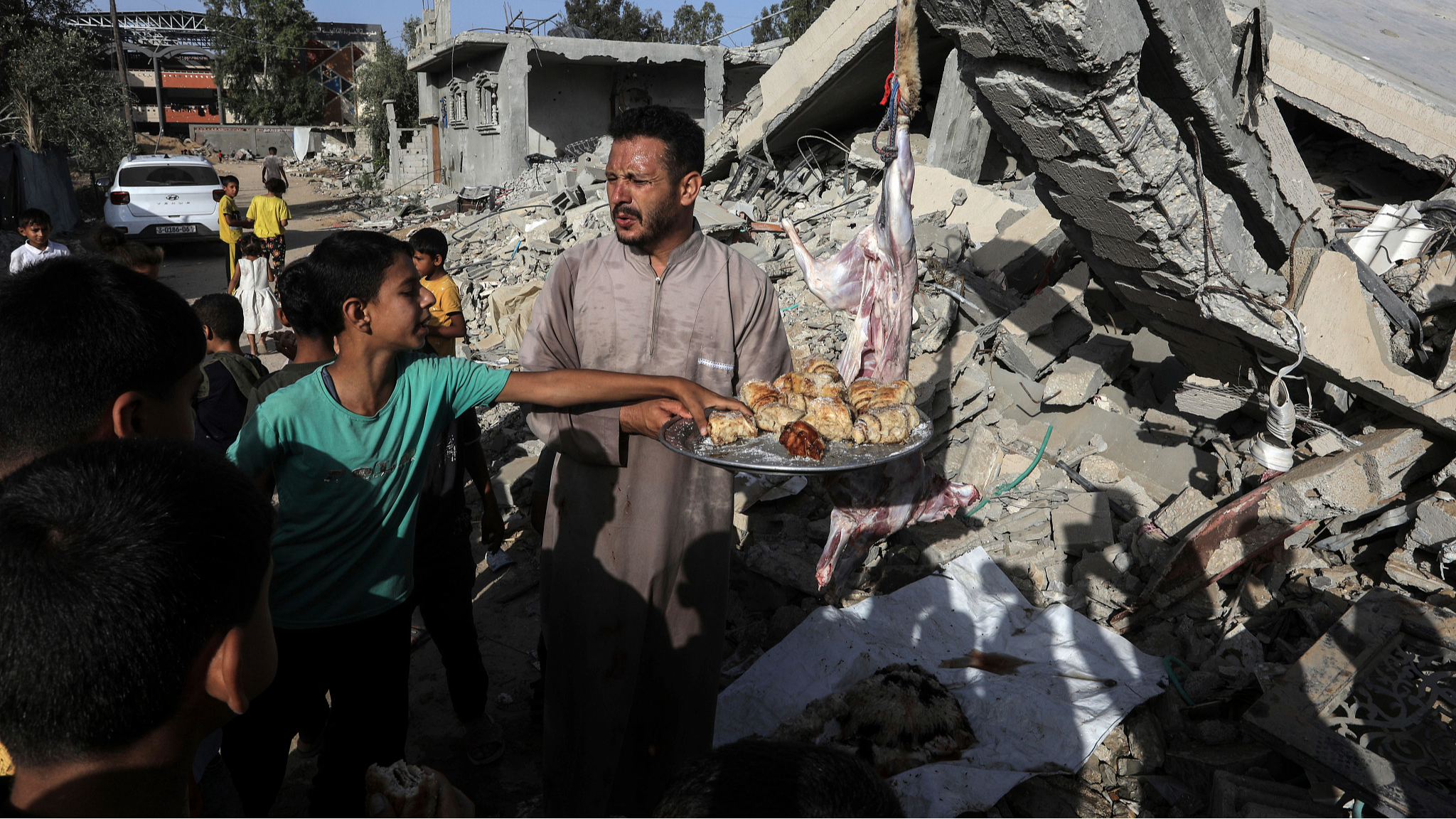 A Palestinian man distributes sweets to people next to the rubble of his house which was destroyed by Israeli warplanes, on the first day of Eid al-Adha, in the city of Khan Yunis in the southern Gaza Strip, June 16, 2024. /CFP