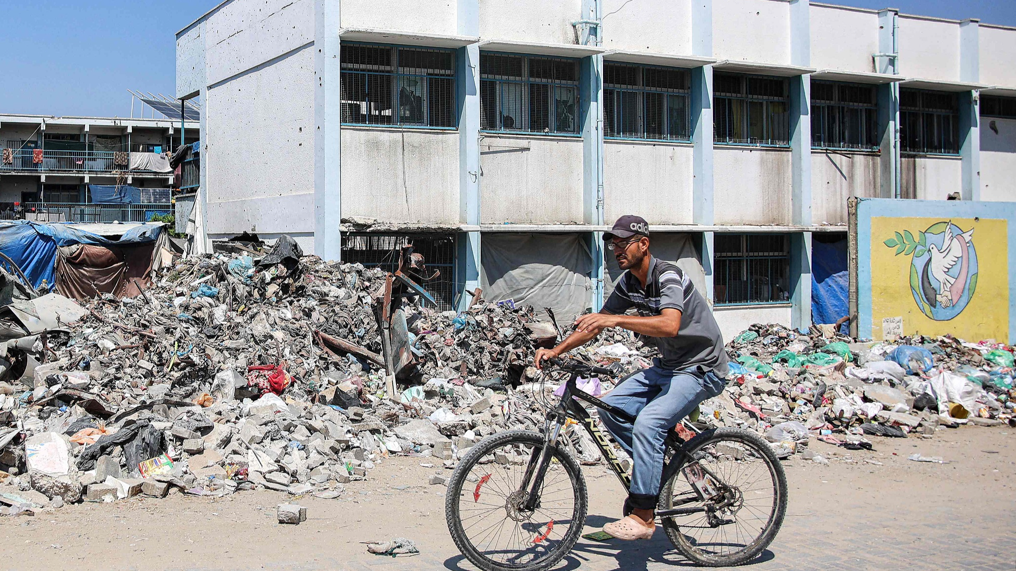 A man cycles past trash and rubble outside a school run by the UN Relief and Works Agency for Palestine Refugees, being used as a makeshift shelter for Palestinians displaced by conflict, in the Jabalia camp for Palestinian refugees in the northern Gaza Strip, August 14, 2024. /CFP