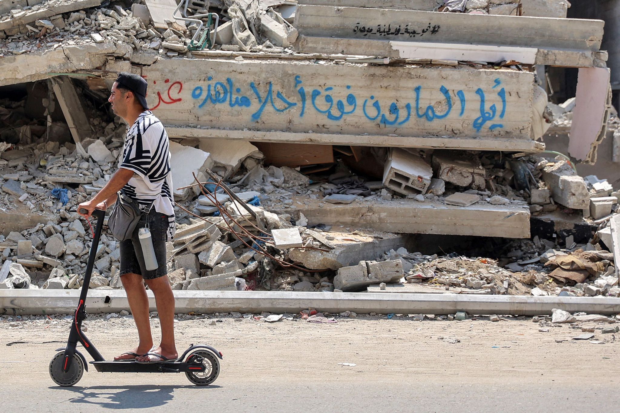 A man rides a scooter past a collapsed building, with a concrete block bearing graffiti reading in Arabic 