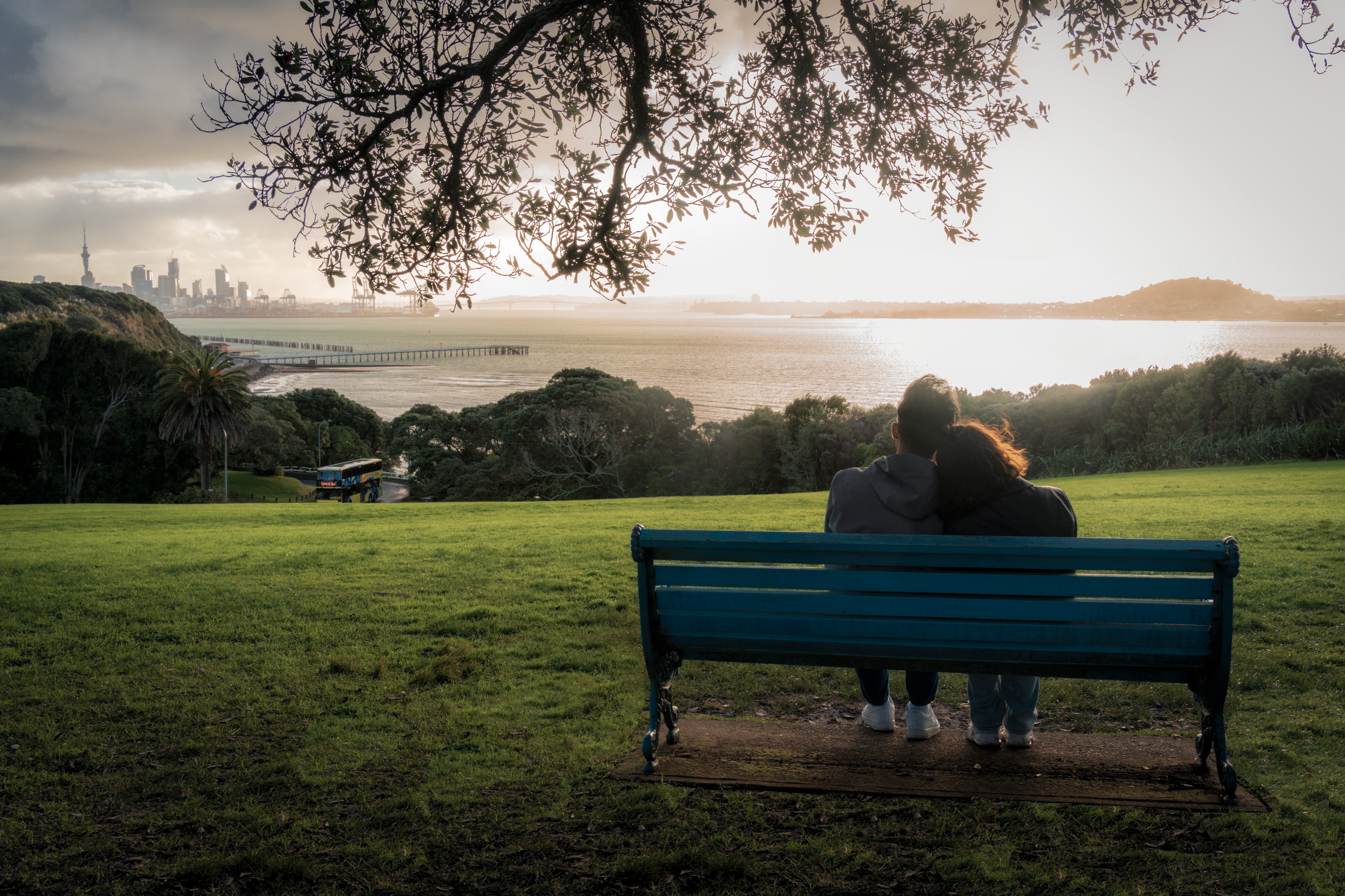 Living in Harmony, Protecting What Is Good: On the green space in Auckland, New Zealand, the sunset casts a gentle glow over the city and the ocean. A couple leans quietly against a bench; lush vegetation, clear waters, and the urban skyline blend harmoniously, while sunlight filters through the clouds and spills down.
This scene aligns with the UN Sustainable Development Goals of 