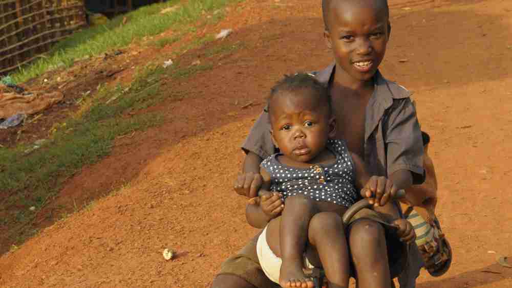A child rides a tricycle with his young brother aboard in the slums of Kampala, Uganda, August 24, 2023. The sun beats down as they push forward against the weight of poverty. This scene highlights the urgency of poverty eradication, aligning with the UN Sustainable Development Goals of 