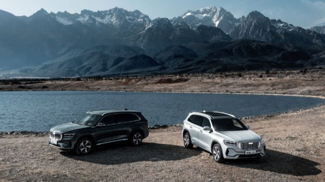 Two Geely Xingyue L cars are parked by a serene lake, with mountain peaks rising in the background. /Geely