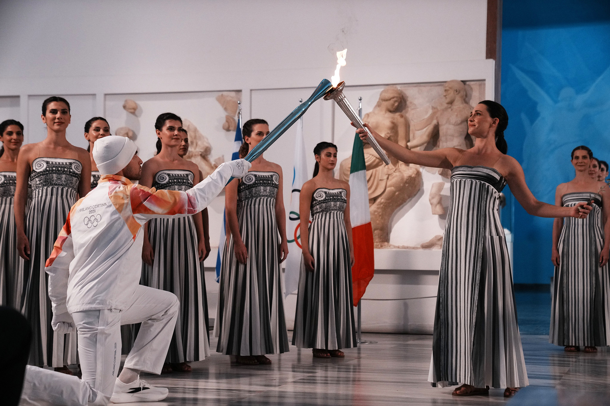 Actress Mary Mina (R) plays the role of a high priestess passing the Olympic flame to the first torchbearer, Greek rower Petros Gaidatzis, during the flame lighting ceremony for the Milano Cortina 2026 Winter Olympics at the archaeological museum in Olympia, Greece, November 26, 2025. /VCG