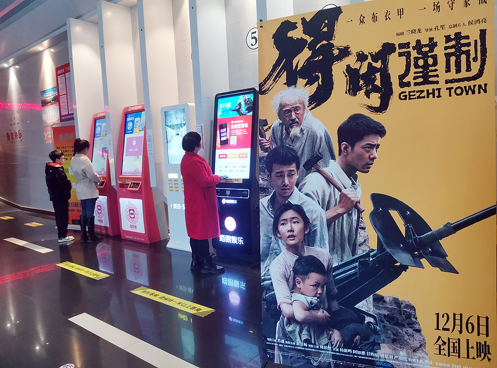 Moviegoers purchase tickets at self-service kiosks beside a poster for 