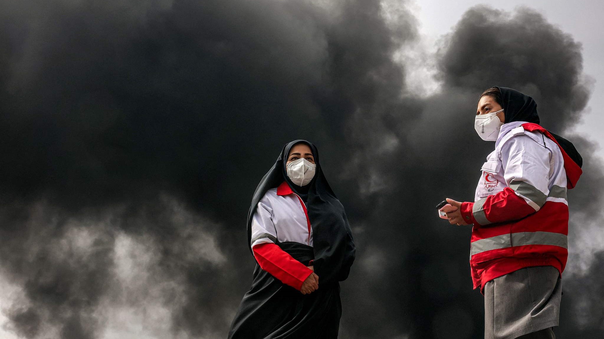 Women members of Iran's Red Crescent society stand near smoke plumes from an ongoing fire following an overnight airstrike on the Shahran oil refinery, northwestern Tehran, March 8, 2026. /VCG