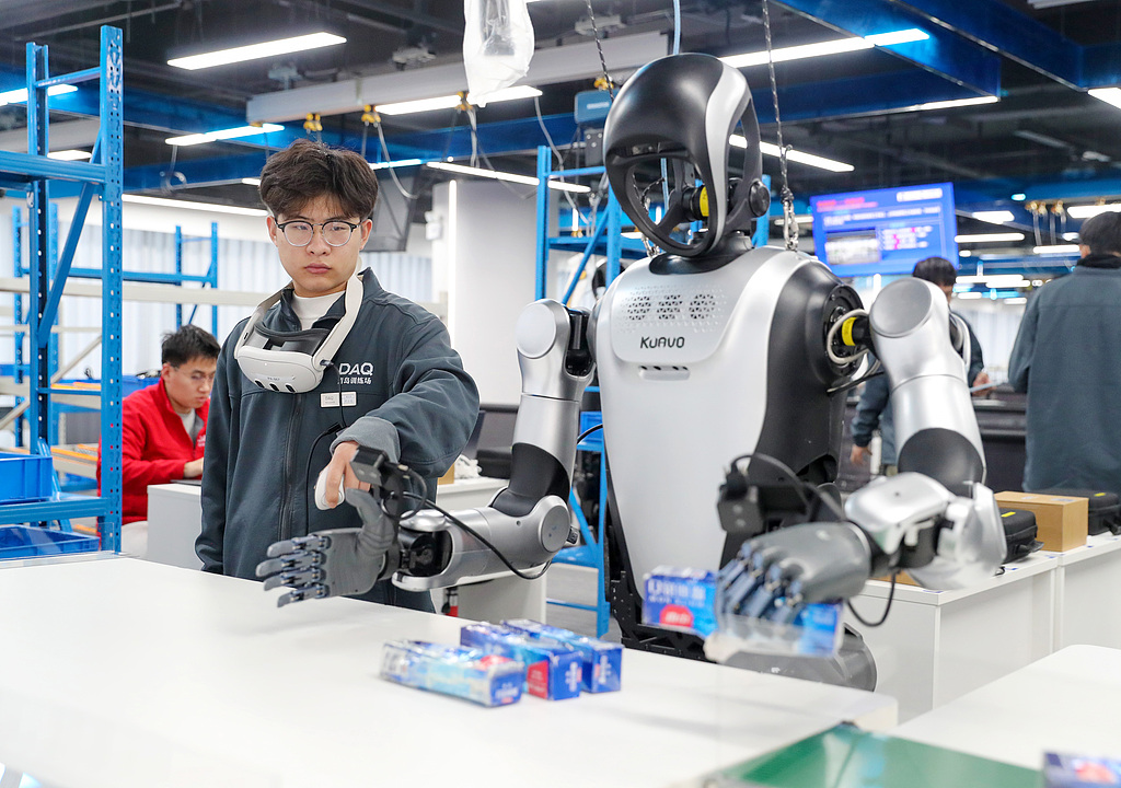 A worker guides a humanoid robot to load toothpaste at a data collection and training facility in Qingdao City, Shandong Province, China, March 18, 2026./ VCG