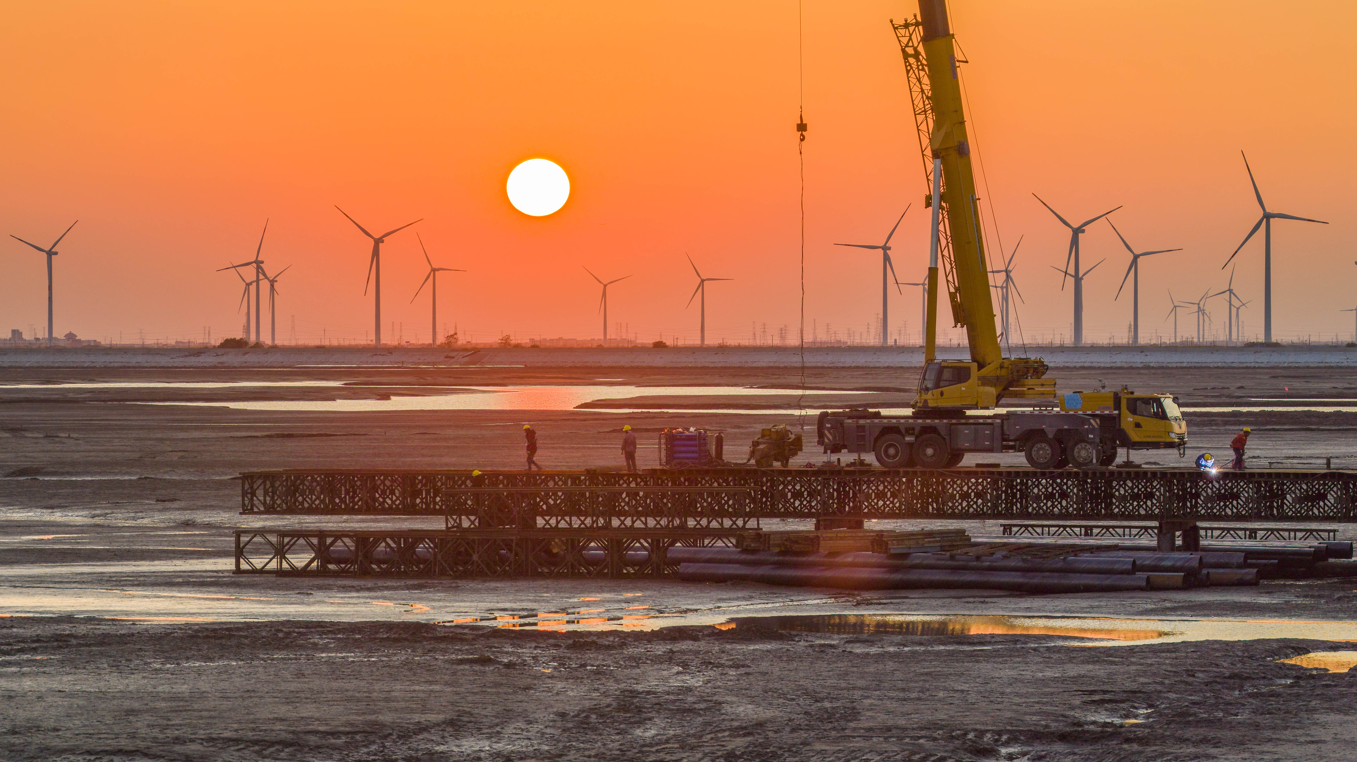 Workers constructing a Bailey bridge for a 500 kV transmission line project of the 2×1,000 MW large-scale High-Efficiency Low Emissions (HELE) coal-fired power plant in Nantong, with wind turbine arrays seen in the background. Nantong, Jiangsu Province, October 28, 2025. /VCG