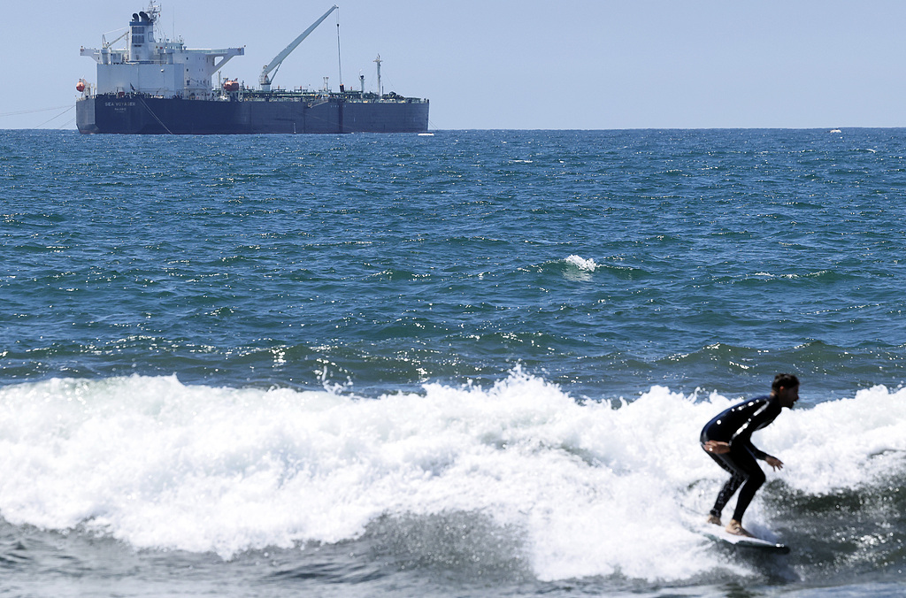 A surfer rides a wave in front of the Sea Voyager crude oil tanker, anchored in the Pacific Ocean off the coast of the Chevron El Segundo refinery in El Segundo, California, US, April 23, 2026. /VCG