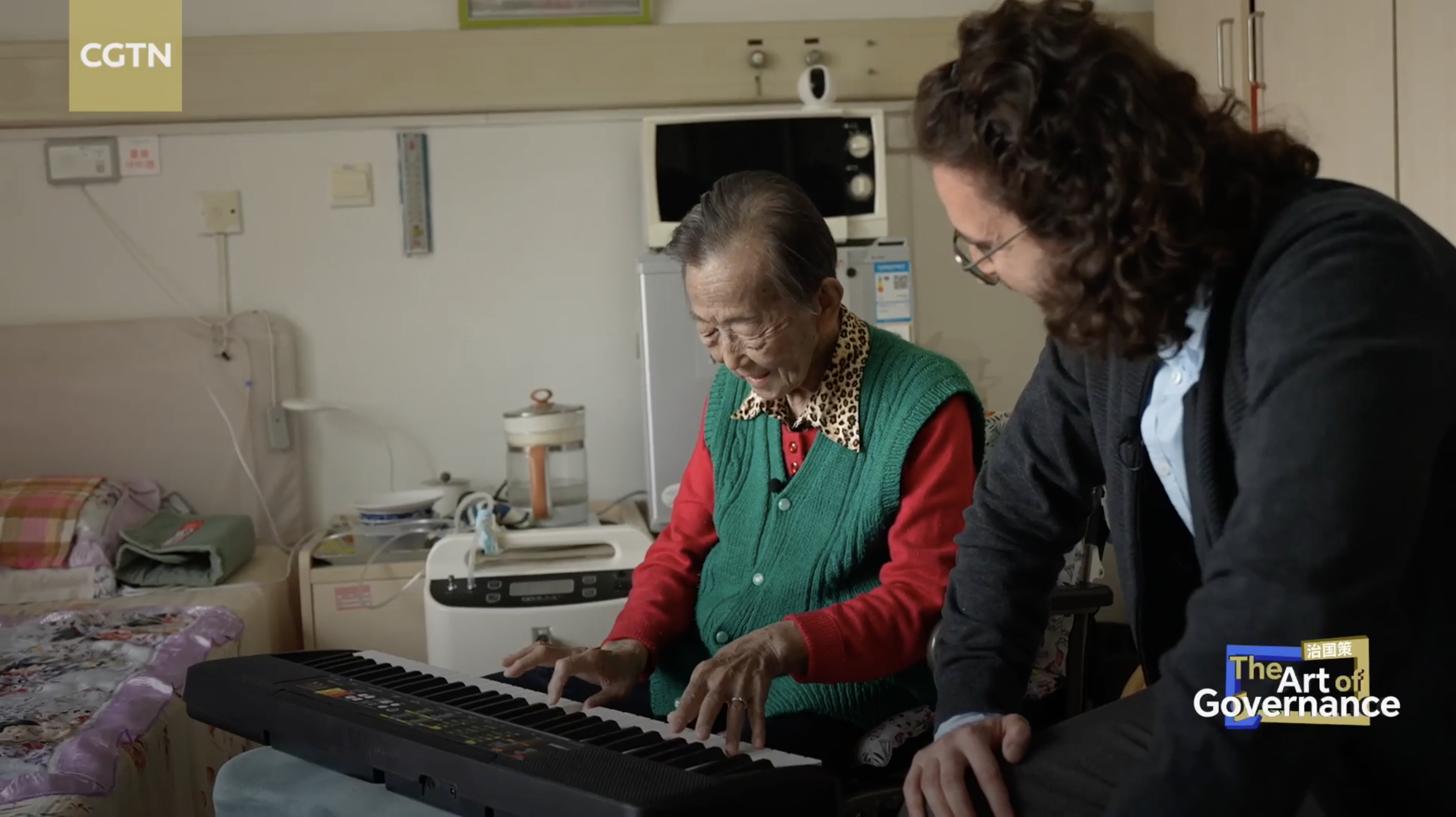Yu Suqin, an 88-year-old resident in Beijing's Xicheng District, plays the piano in his room while a CGTN reporter, Nadim Diab, looks on. /CGTN
