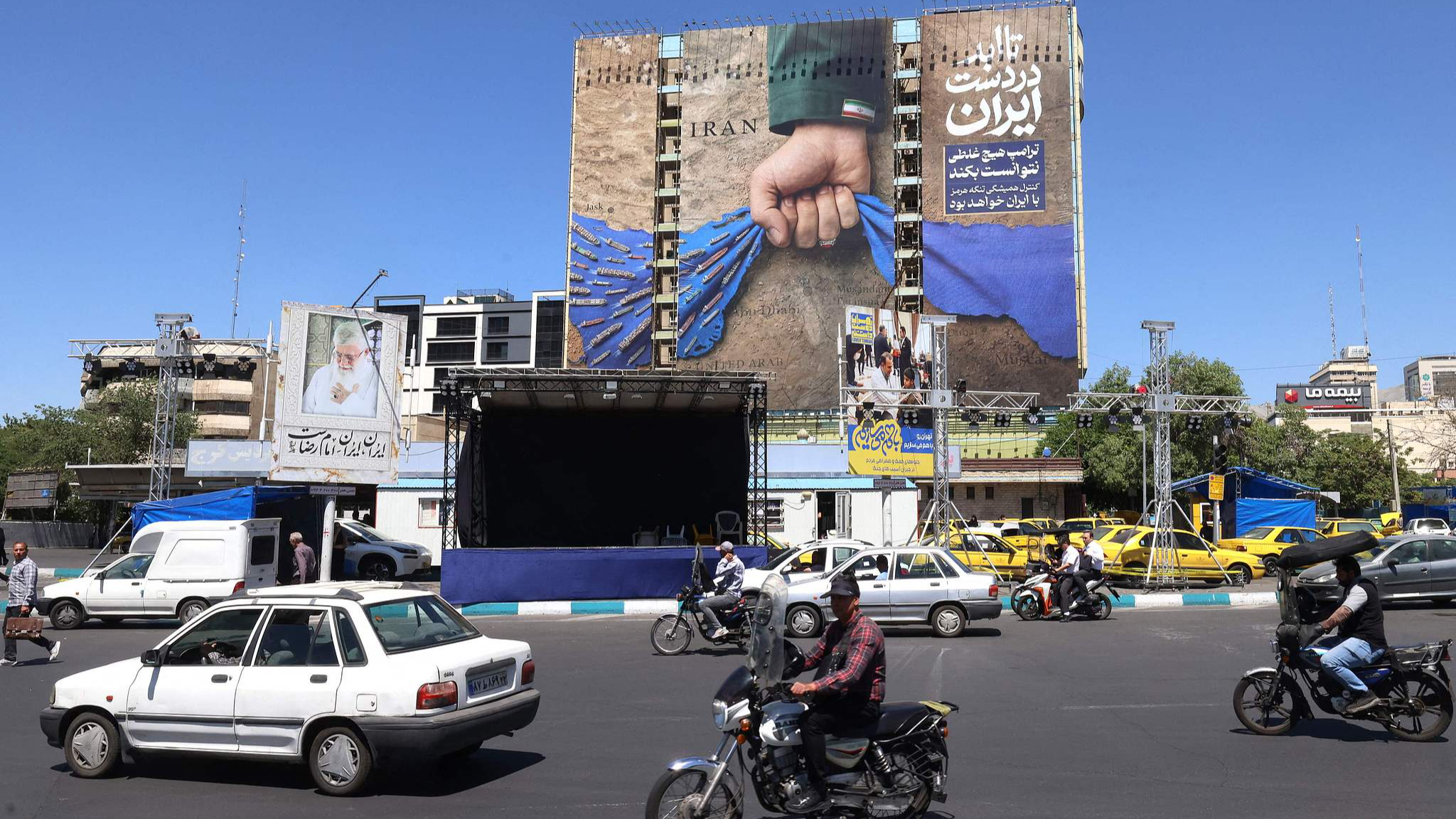 Vehicles drive past a billboard on the facade of a building depicting the Strait of Hormuz, with a Persian caption reading 