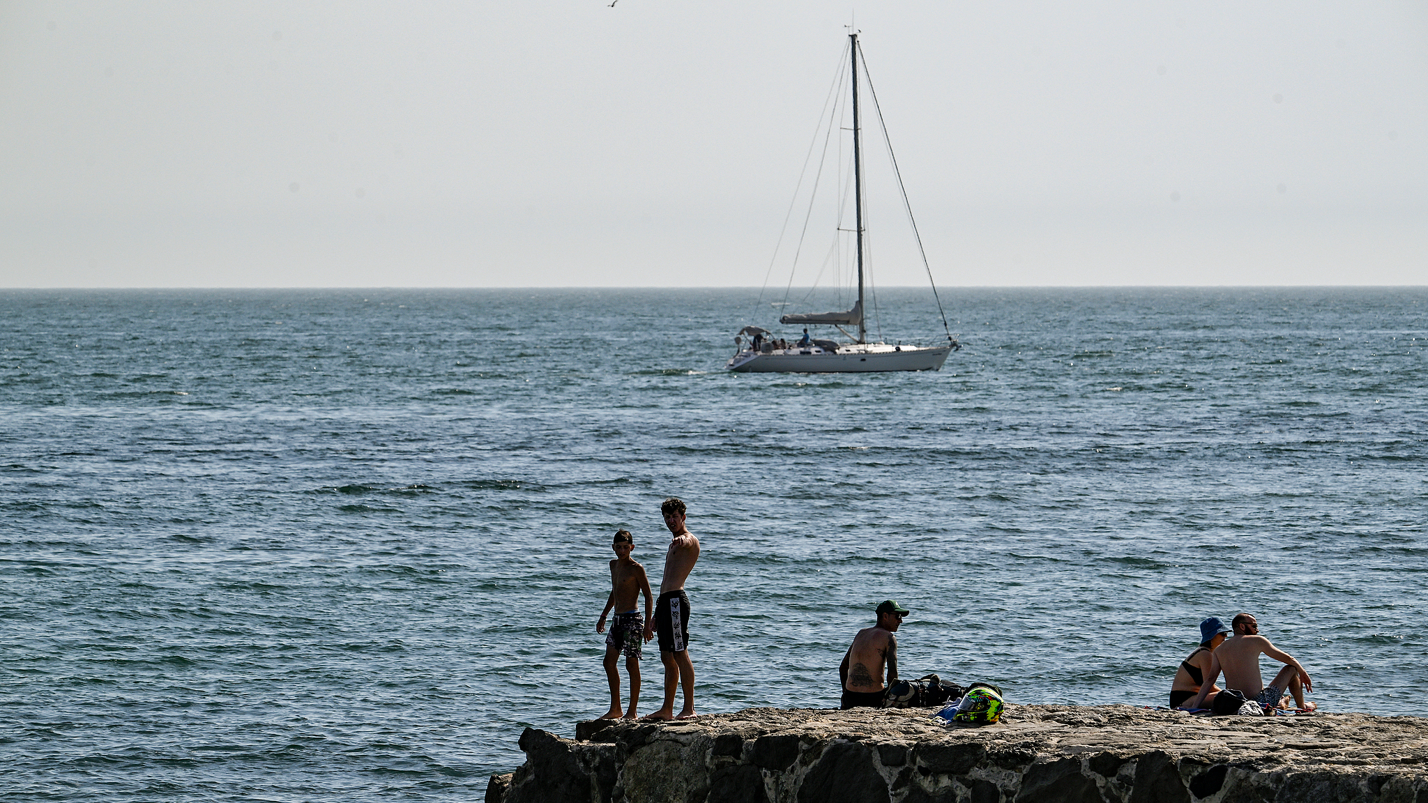 Youngsters stand at the end of the wave breaker as beachgoers crowd Carcavelos beach as temperatures reach 37 degrees Celsius during a sunny afternoon on July 05, 2026 in Carcavelos, Portugal. /VCG