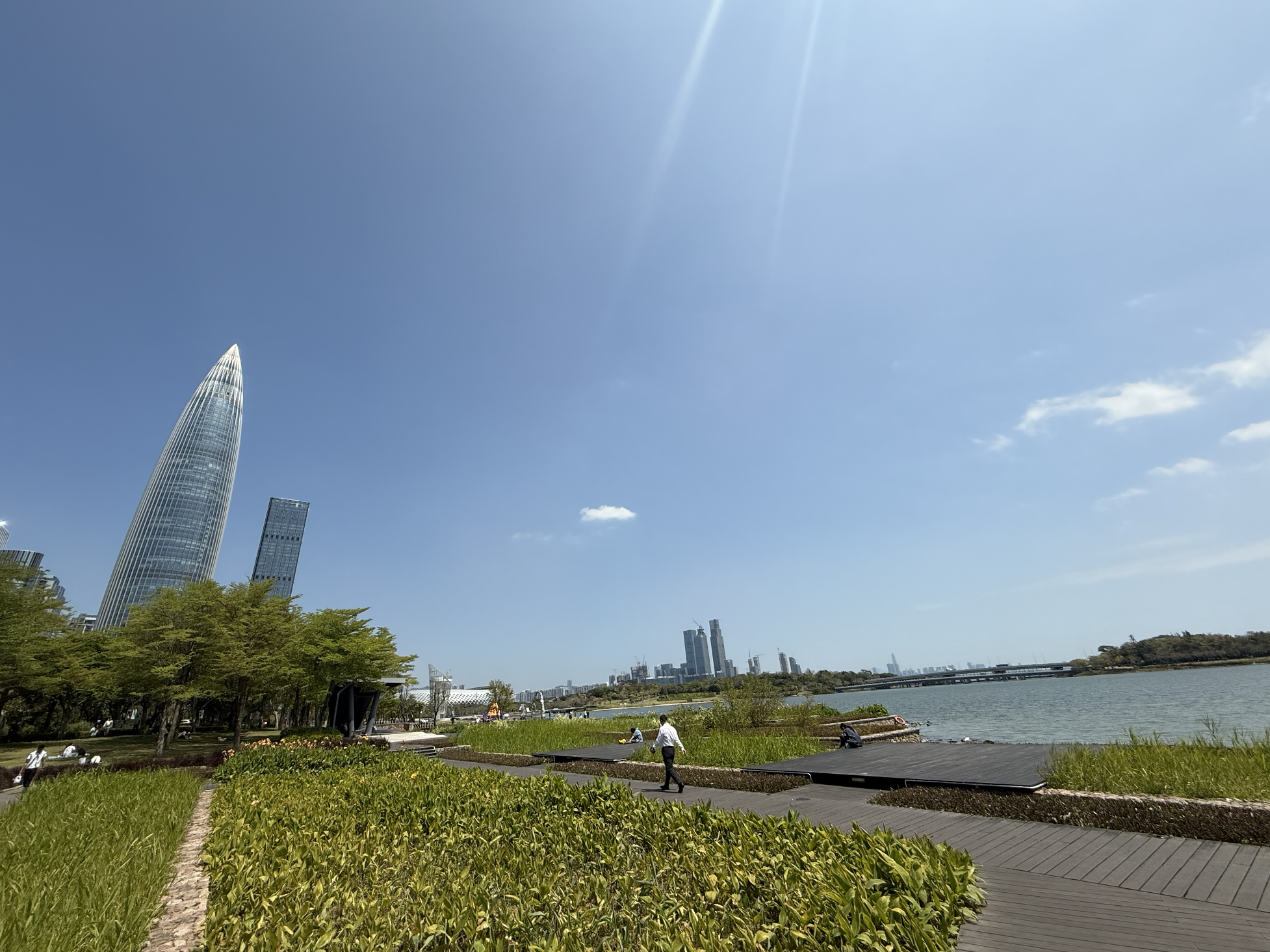 Shenzhen Talent Park, Nanshan, the Spring Bamboo tower catching the afternoon light above the park's lakeside walkway, with the wider Nanshan skyline stretching along Shenzhen Bay in the distance. 
