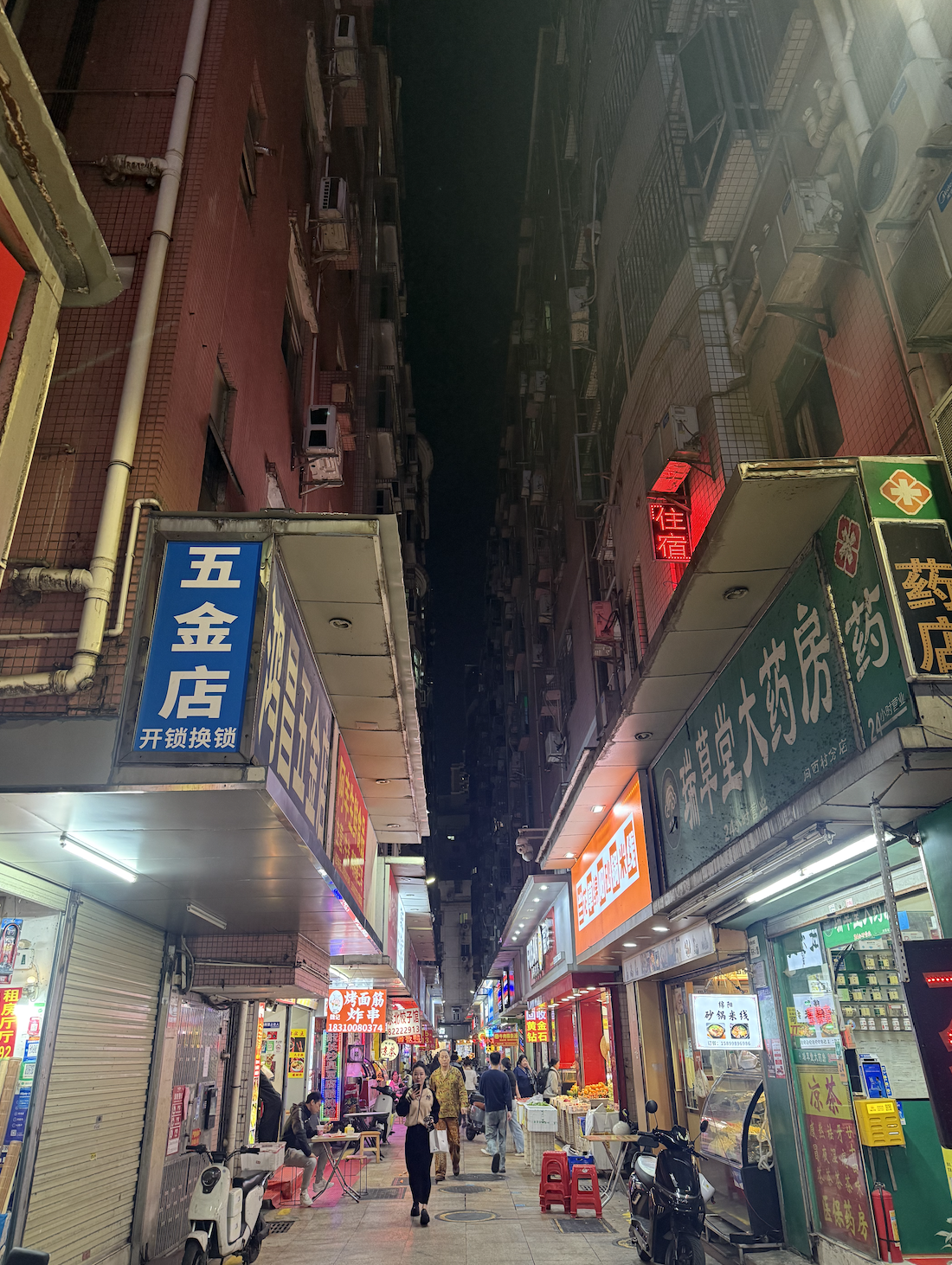 Ground level inside Xiangxicun urban village, with hardware stores, pharmacies, a lodging sign glowing red, motorbikes parked.
