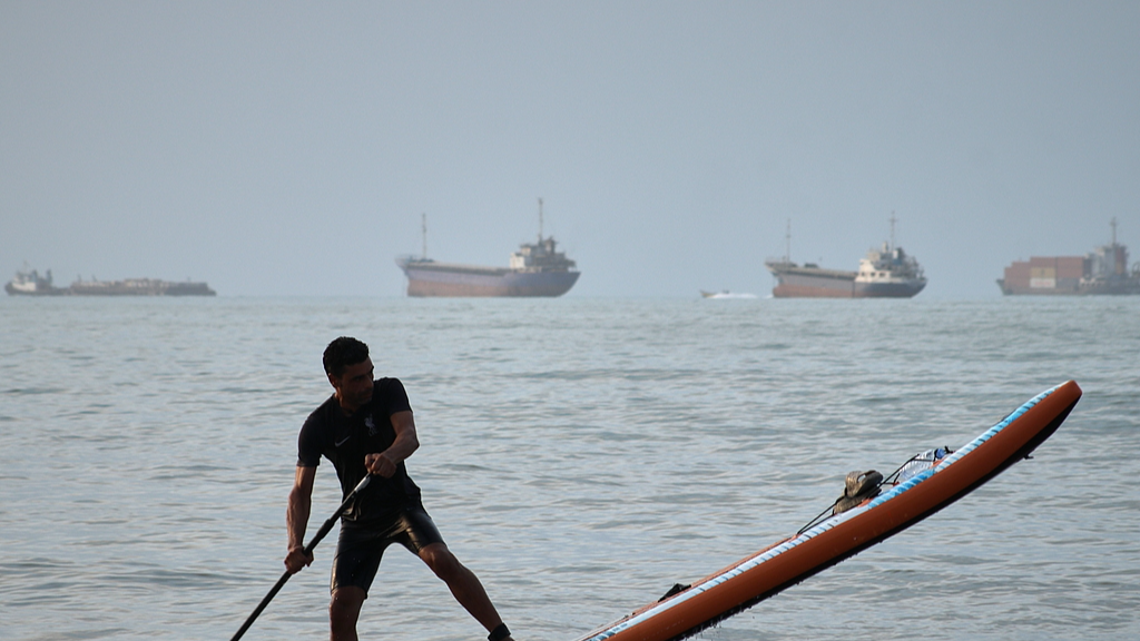 A man paddles a stand-up paddleboard as cargo ships and other commercial vessels appear anchored in the Strait of Hormuz off Bandar Abbas, Iran, July 27, 2026. /VCG