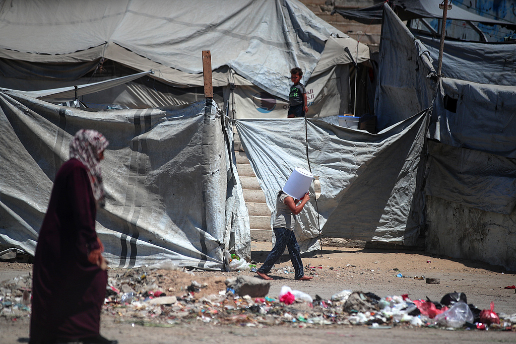 A displaced Palestinian girl walks past her family's tent with a plastic container balanced on her head to shield herself from the sun in Gaza City, August 2, 2026. /VCG