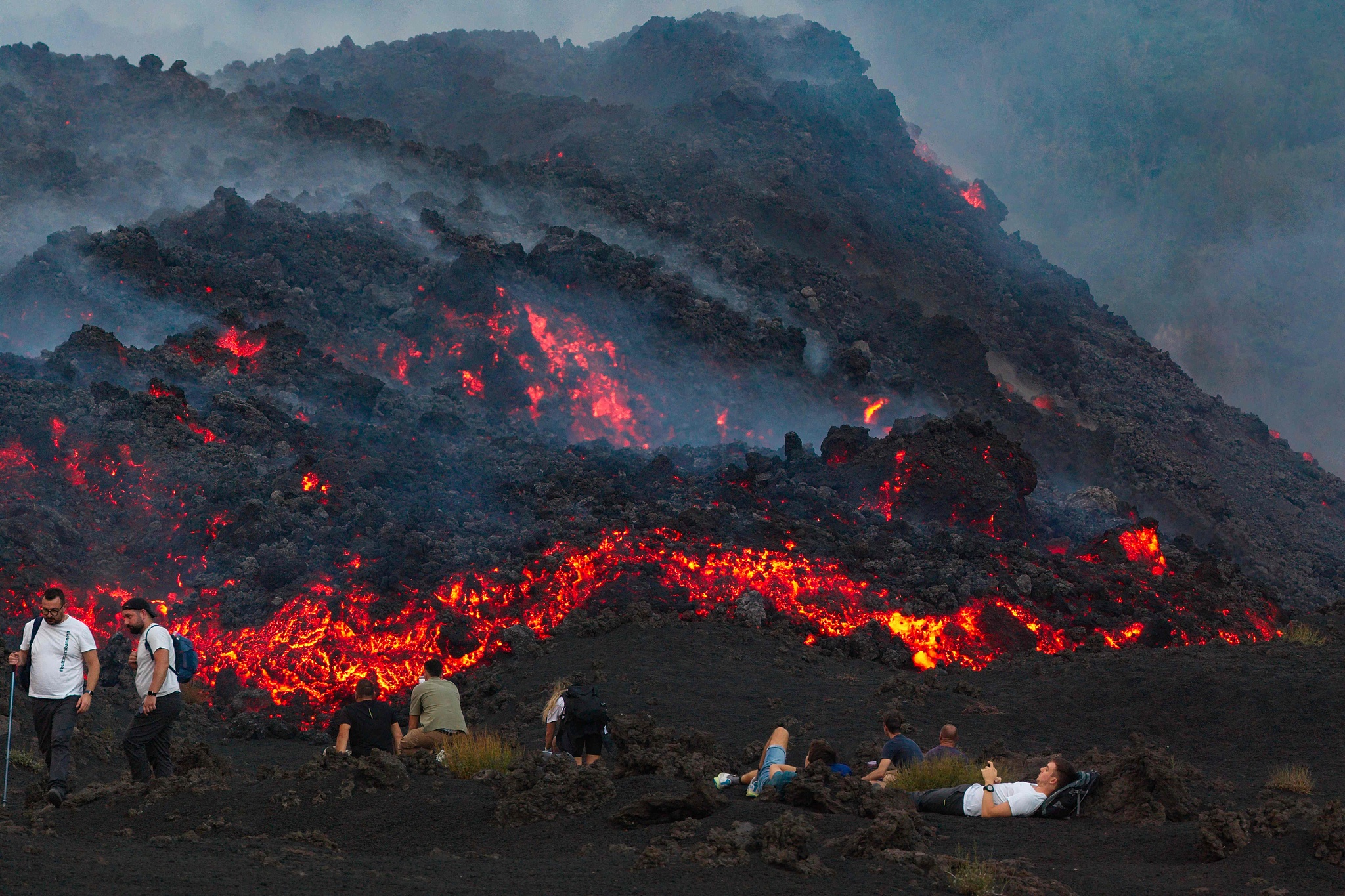A photo shows visitors sitting next to lava flowing from Mount Etna volcano during an eruption, in Catania, in the Mediterranean island of Sicily, Italy, August 12, 2026. /VCG