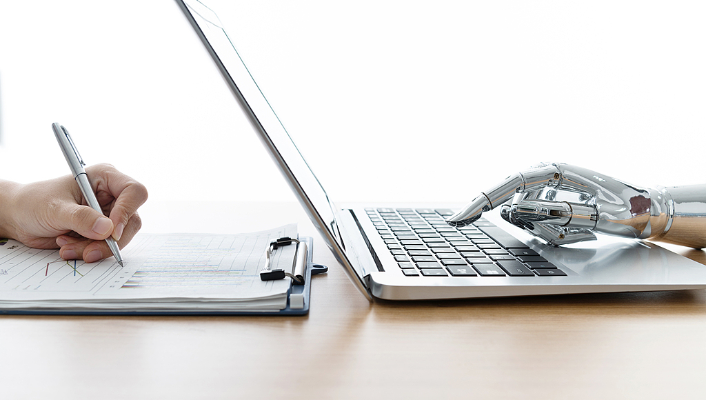 An illustration shows a robotic hand using a laptop as a man writes by hand. /VCG